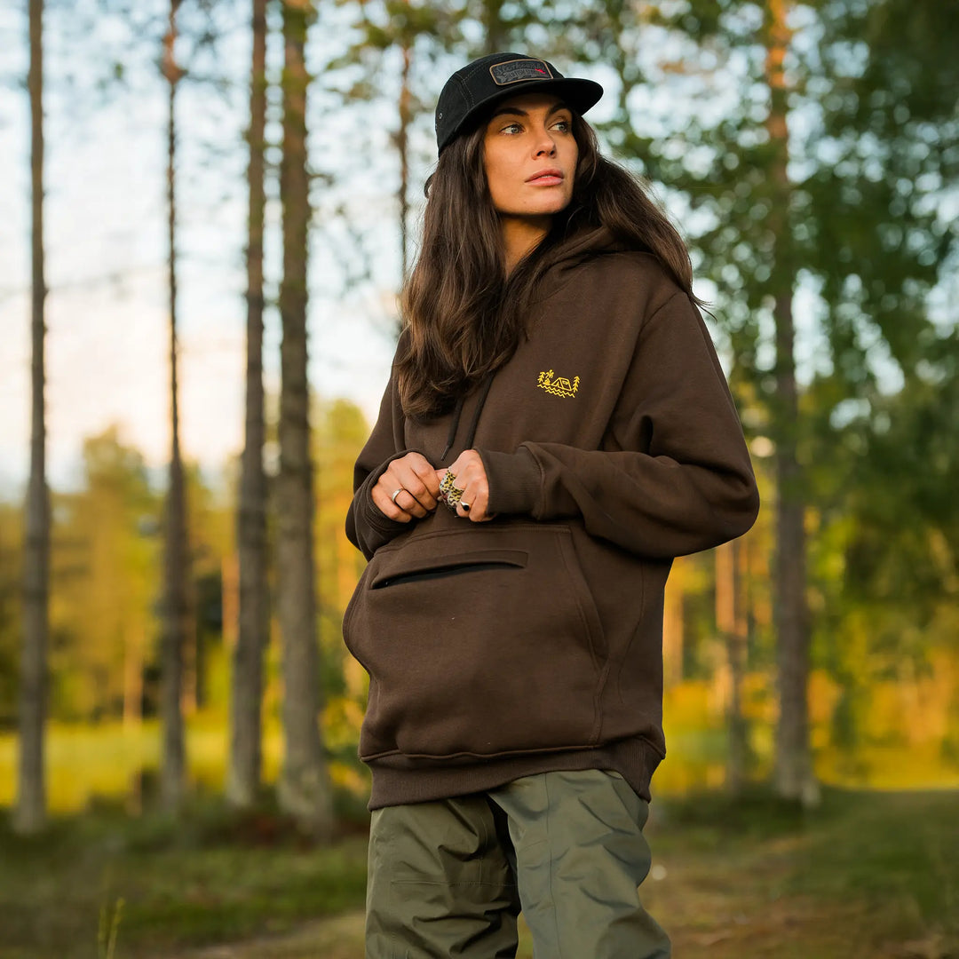 Bluza wędkarska Wild Fish Stories El Clasico noszona przez kobietę w lesie, brązowa bluza outdoorowa z kapturem i minimalistycznym logo