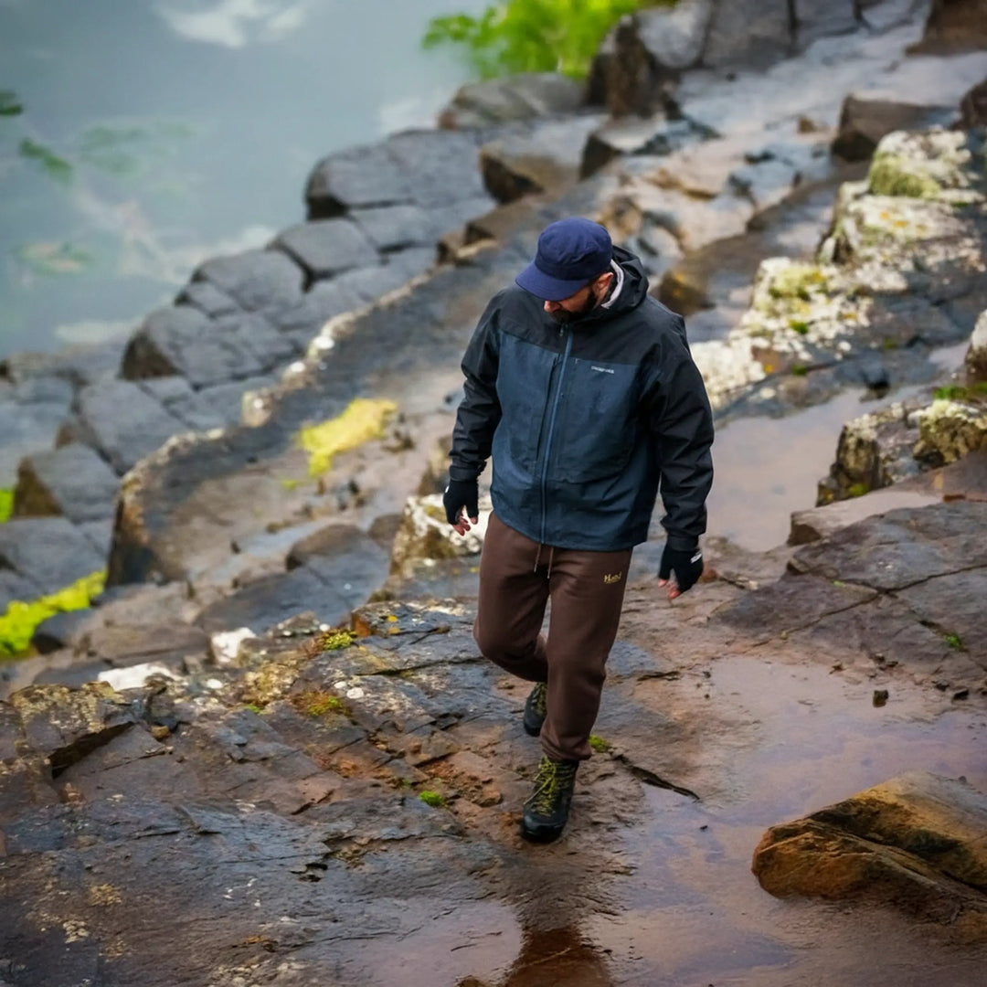 Spodnie wędkarskie El Clasico Wild Fish Stories - wędkarz w brązowych spodniach outdoor nad rzeką kamienisty brzeg