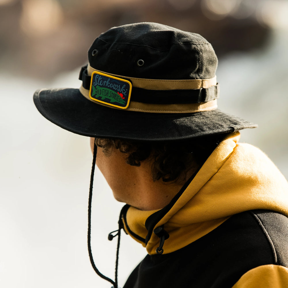 Kapelusz Angler Sterkowski x Wild Fish Stories noszony nad wodą, woskowana bawełna British Millerain, kapelusz outdoorowy i wędkarski z paskiem i sznurkiem pod brodą, przeznaczony na wyprawy w teren