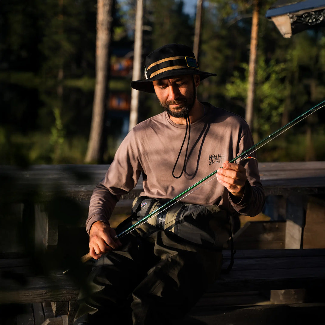 Longsleeve Wild Fish Stories Camper Chill noszony nad wodą, mężczyzna przygotowujący wędkę, outdoorowy longsleeve z logo na piersi, klimat kamper, slow fishing i wypoczynek w naturze