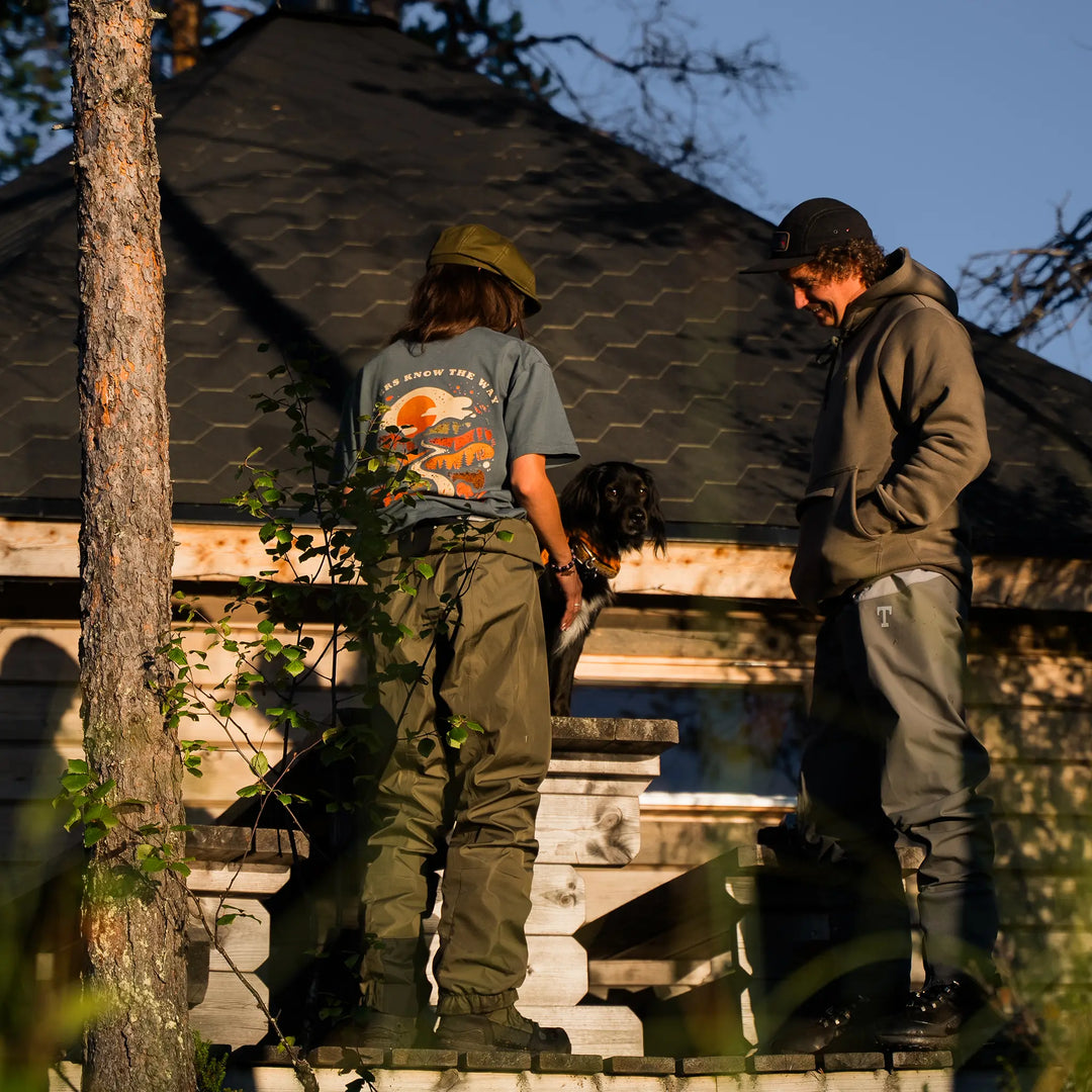 Koszulka Wild Fish Stories z grafiką Rivers Know the Way noszona podczas outdoorowego wypoczynku, ludzie i pies przy drewnianej chacie w naturze