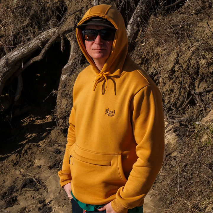 Man wearing Wild Fish Stories mustard yellow hoodie with chest logo, standing outdoors in a natural rocky setting
