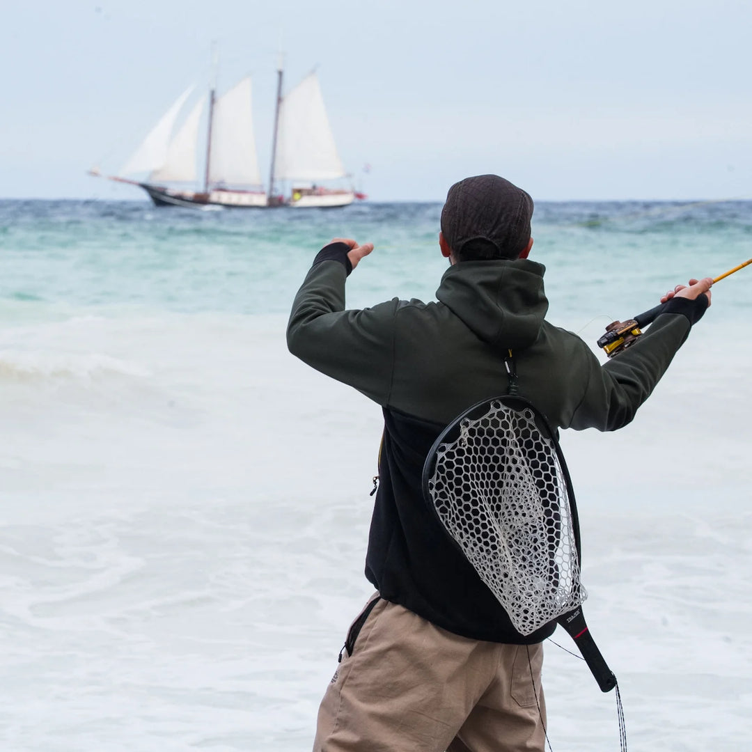 Angler in Wild Fish Stories olive and black hoodie casting a fly rod on the beach with a tall ship sailing in the background