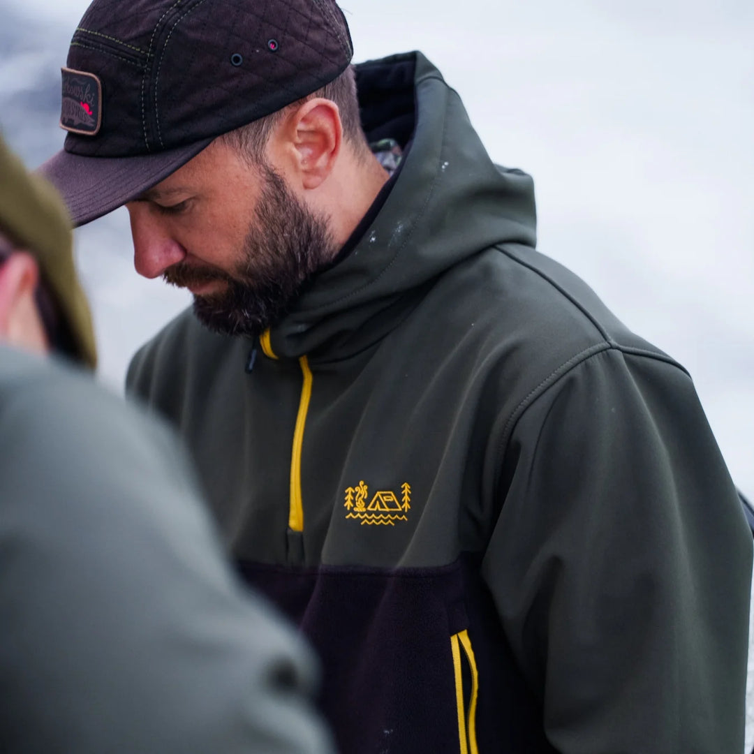 Close-up of Wild Fish Stories olive and black hoodie with embroidered gold logo on chest, worn by bearded angler outdoors