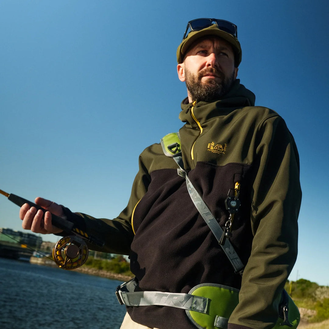 Angler wearing Wild Fish Stories olive and black hoodie with yellow accents, holding a fly fishing rod by the water on a sunny day