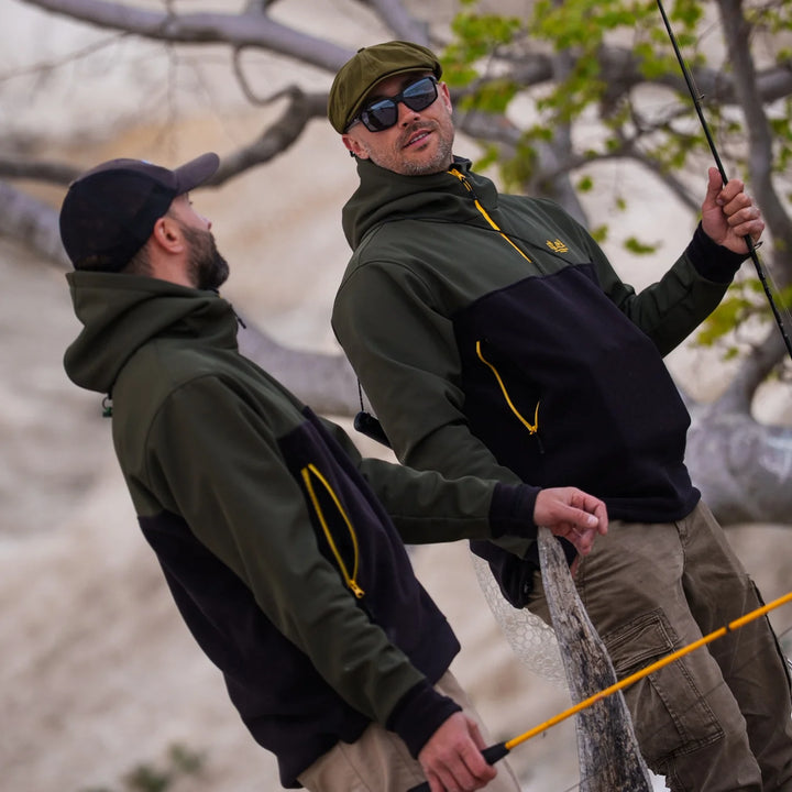 Two anglers wearing Wild Fish Stories olive and black hoodies with yellow zippers, fishing together outdoors with rods and landing net