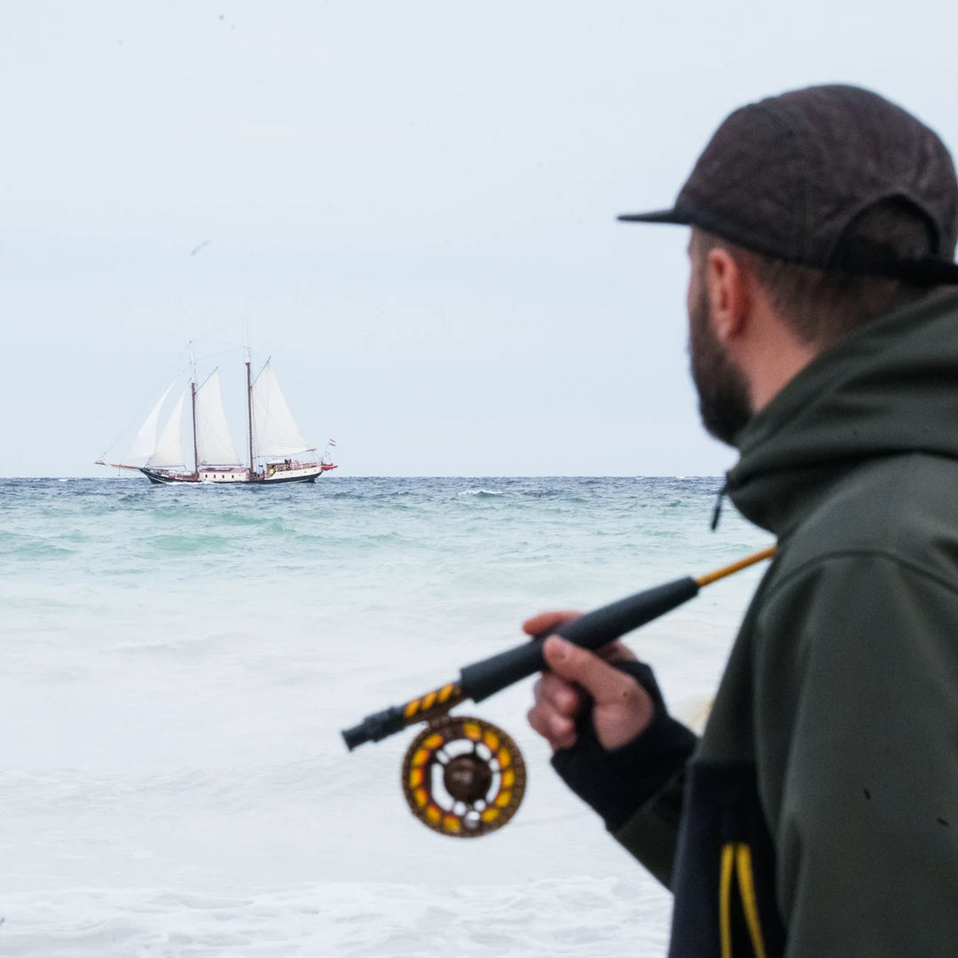 Angler in Wild Fish Stories olive hoodie holding a fly reel on the beach, watching a tall ship sailing on the sea