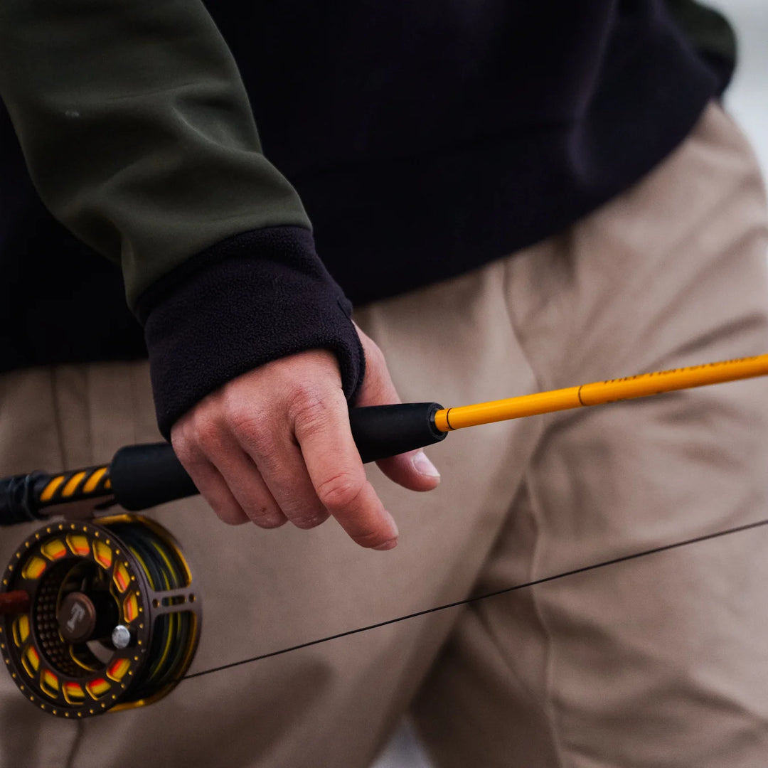 Close-up of angler's hand gripping a fly rod with a gold fly reel, wearing Wild Fish Stories olive and black hoodie sleeve